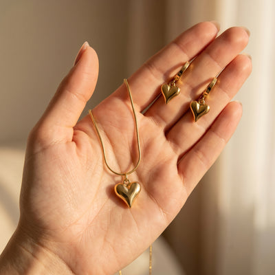 Hand holding gold heart-shaped jewelry set against a neutral background