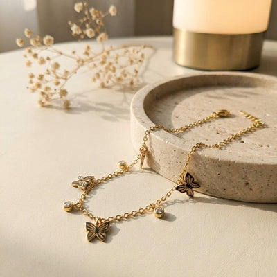Gold butterfly necklace on a stone surface with dried flowers and a lamp in the background