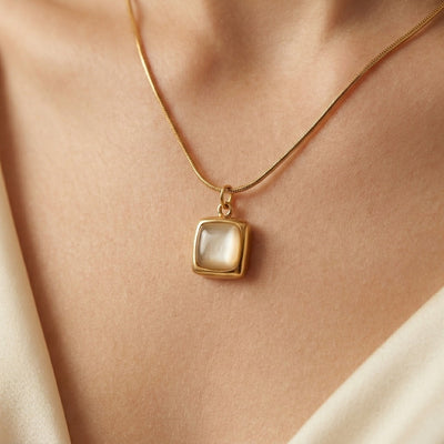 Gold necklace with a square pendant on a person wearing a beige top.